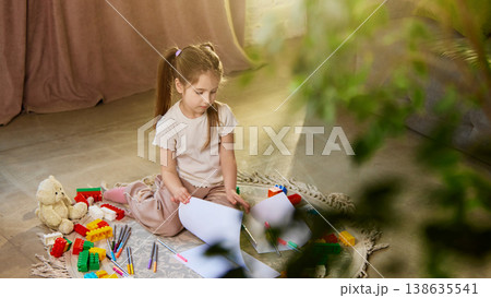 Little girl sorting drawings and papers sitting on floor with toys. 138635541