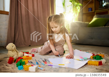 Little girl drawing on paper on floor with colorful toys in warm sunlight. Little girl drawing on paper on floor with colorful toys in warm sunlight. 138635549