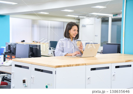 オフィスのスタンディングデスクでコーヒーを持ちながらパソコン作業をする女性 138635646