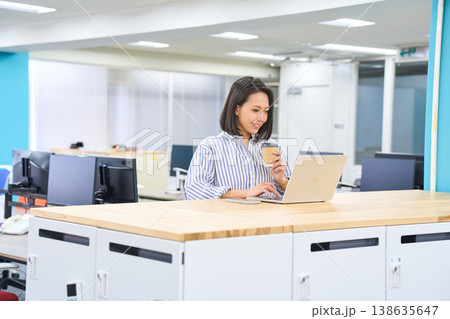 オフィスのスタンディングデスクでコーヒーを持ちながらパソコン作業をする女性 オフィスのスタンディングデスクでコーヒーを持ちながらパソコン作業をする女性 138635647
