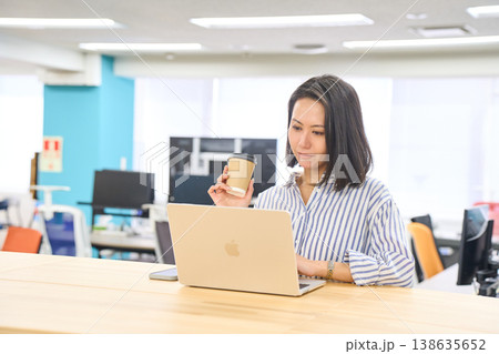 オフィスのスタンディングデスクでコーヒーを持ちながらパソコン作業をする女性 138635652