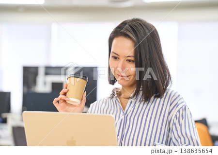 オフィスのスタンディングデスクでコーヒーを持ちながらパソコン作業をする女性 オフィスのスタンディングデスクでコーヒーを持ちながらパソコン作業をする女性 138635654