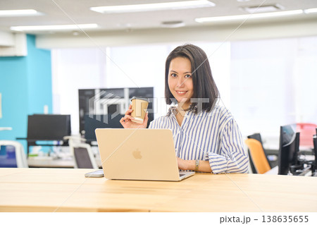 オフィスのスタンディングデスクでコーヒーを持ちながらパソコン作業をする女性 オフィスのスタンディングデスクでコーヒーを持ちながらパソコン作業をする女性 138635655