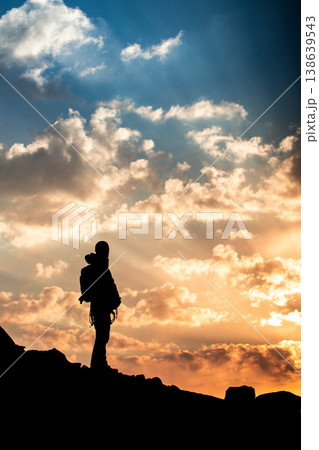 Vertical shot of a climber silhouette on a mountain peak during a bright golden sunset with blue sky and white clouds. 138639543