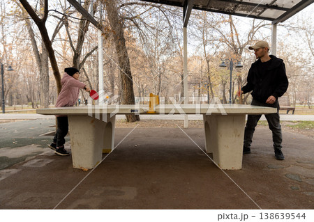 Full side view of a man and a girl playing ping pong on a concrete table surrounded by trees in a quiet outdoor setting. Full side view of a man and a girl playing ping pong on a concrete table surrounded by trees in a quiet outdoor setting. 138639544