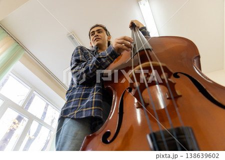 Dynamic low angle view of a young college student playing a large wooden double bass in a bright music room. Dynamic low angle view of a young college student playing a large wooden double bass in a bright music room. 138639602