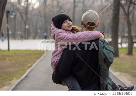 A father walks through a park carrying his daughter in a pink coat, who is hugging him from behind and smiling. 138639603