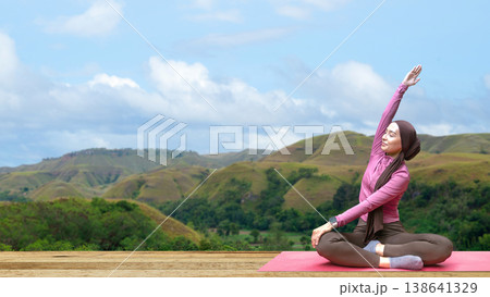 A young Indonesian southeast asian muslim woman in a hijab and modest sportswear practices yoga and stretching on a red mat with a stunning mountain landscape background A young Indonesian southeast asian muslim woman in a hijab and modest sportswear practices yoga and stretching on a red mat with a stunning mountain landscape background 138641329