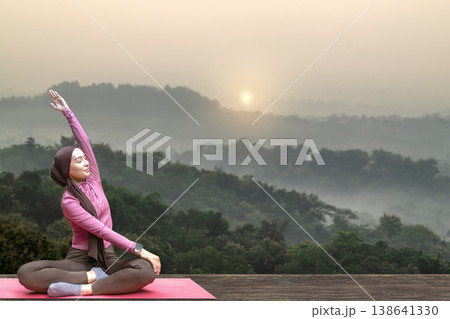 A young Indonesian southeast asian muslim woman in a hijab practices yoga and stretching on a mat outdoors against a beautiful misty mountain landscape during sunrise. A young Indonesian southeast asian muslim woman in a hijab practices yoga and stretching on a mat outdoors against a beautiful misty mountain landscape during sunrise. 138641330