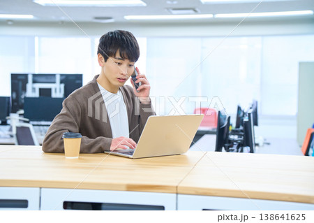 フリーアドレスデスクでパソコンを開きスマホで通話する男性社員 フリーアドレスデスクでパソコンを開きスマホで通話する男性社員 138641625