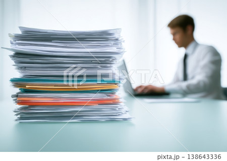 Person handling pressing documents next to a laptop. Busy office worker typing on keyboard beside towering pile of paperwork on desk. Stressed professional man working under pressure amidst deadline 138643336