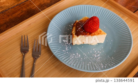 close up of a slice of basque cheesecake with a strawberry on top placed on a blue plate with white sugar powder on a wooden tray with two small forks 138644591