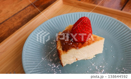 close up of a slice of basque cheesecake with a strawberry on top placed on a blue plate with white sugar powder on a wooden tray with two small forks 138644592