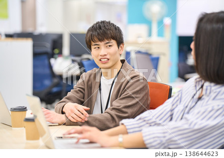 フィスのデスクで同僚の女性と笑顔で会話する若い男性社員 138644623