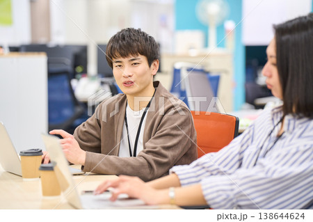 フィスのデスクで同僚の女性と笑顔で会話する若い男性社員 138644624