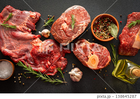 Several types of beef steak are laid out on a dark surface with fresh herbs and spices. Ingredients include garlic, pepper, and oil, showing preparation for cooking in a kitchen Several types of beef steak are laid out on a dark surface with fresh herbs and spices. Ingredients include garlic, pepper, and oil, showing preparation for cooking in a kitchen 138645287