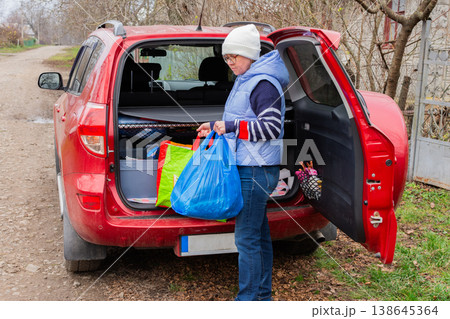 A woman stands by her red car on a dirt road. She holds a blue bag and lifts items from her car after visiting a market close to her home A woman stands by her red car on a dirt road. She holds a blue bag and lifts items from her car after visiting a market close to her home 138645364