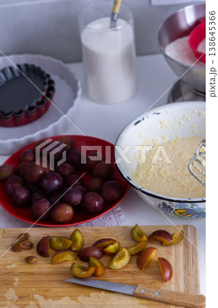 Cook is preparing to make plum pie in a home kitchen. Ingredients like plums, flour, sugar, and butter are on the counter. Tools for mixing and baking are nearby 138645366