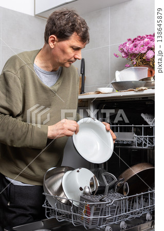 A man stands in his kitchen taking clean dishes out of the dishwasher. He focuses on the process of unloading the clean dishes and organizing them for use 138645879
