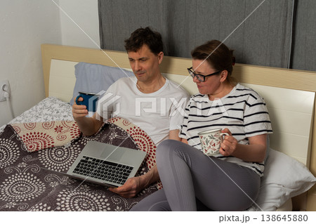 A man and a woman sit together on a bed. They both hold drinks. The man looks at a laptop, while the woman appears to read something on her phone in their home 138645880