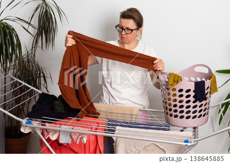 A 50-year-old woman is in her room. She is hanging washed laundry on a drying rack. A basket of clothes is nearby. She looks focused on her task A 50-year-old woman is in her room. She is hanging washed laundry on a drying rack. A basket of clothes is nearby. She looks focused on her task 138645885