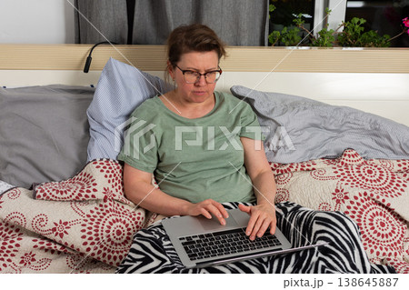A woman sits on bed in bedroom and works on laptop. She has blankets around her and seems focused on task while enjoying her own space 138645887