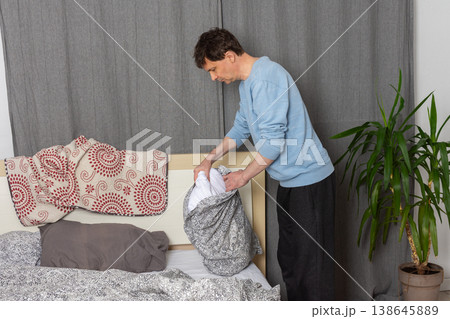 A 53-year-old man replaces the bed linen in his bedroom. He gathers clean sheets and works on making the bed comfortable for sleeping A 53-year-old man replaces the bed linen in his bedroom. He gathers clean sheets and works on making the bed comfortable for sleeping 138645889