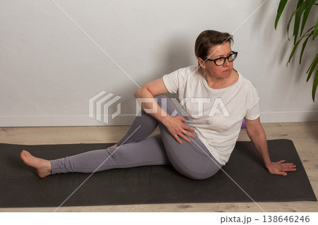 A 52-year-old woman is engaged in an exercise routine at home. She is sitting on a yoga mat and performing a stretching move, focusing on her posture and breathing 138646246