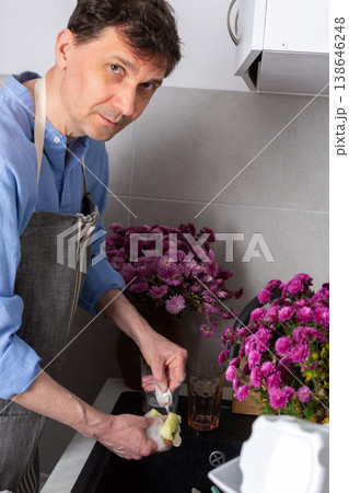 A man stands at a sink in a kitchen, washing dirty dishes and glassware. Flowers sit on the counter as fresh light comes in through the window. The setting is busy and practical 138646248