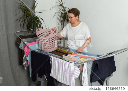 A 52-year-old woman takes freshly cleaned clothes from a drying rack at home. She is focused on her task and handling a basket while surrounded by clothes 138646250