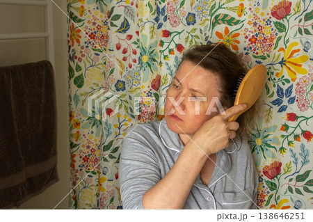 A 50-year-old woman stands in her bathroom holding a wooden hairbrush. She focuses on brushing her hair while facing a floral patterned wall, preparing for her day ahead 138646251