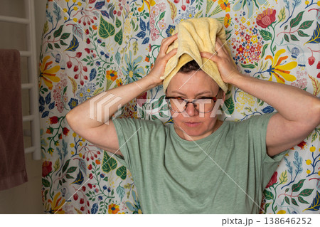 A woman performs her morning bathroom routine. She stands with a towel on her head, looking in the mirror. The colorful floral wall adds brightness to the space A woman performs her morning bathroom routine. She stands with a towel on her head, looking in the mirror. The colorful floral wall adds brightness to the space 138646252