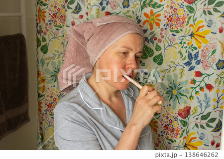 A 50-year-old woman brushes her teeth in a bright bathroom. She wears a towel on her head and sits in front of a floral background. It is morning and she is starting her day 138646262