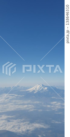 Phone photo of snow-covered Mount Elbrus peeking through clouds, viewed from an airplane window. Aerial mountain scene showing dramatic altitude, winter landscape, and travel perspective. 138646310