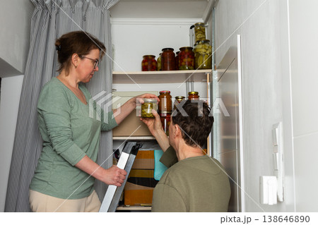 Married couple places jars of canned goods onto shelves in their pantry. They are inside their home with a view of flowers and the city from the balcony. It is autumn Married couple places jars of canned goods onto shelves in their pantry. They are inside their home with a view of flowers and the city from the balcony. It is autumn 138646890