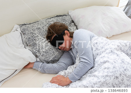 A 50-year-old woman is peacefully sleeping in a cozy bed. She wears a sleep mask and is snuggled under soft blankets, creating a serene atmosphere for rest 138646893