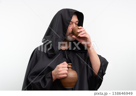 Bearded mature monk with hood drinking beverage from ceramic cup in studio. Bearded mature monk with hood drinking beverage from ceramic cup in studio. 138646949
