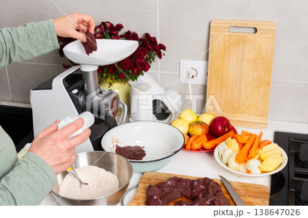 A woman is making liver cutlets in a city kitchen. She is using a grinder to prepare the meat. The kitchen has flowers on the balcony and autumn colors outside A woman is making liver cutlets in a city kitchen. She is using a grinder to prepare the meat. The kitchen has flowers on the balcony and autumn colors outside 138647026