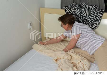 A 53-year-old woman is replacing old bed linen with new ones in her bedroom. She carefully adjusts the fabric as she prepares the bed for use. A cozy blanket is draped on the headboard 138647042