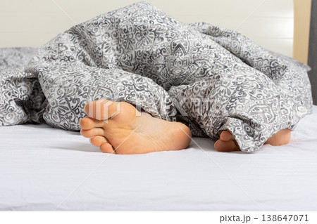 A man enjoys his time relaxing on a bed. His feet can be seen sticking out from under a gray blanket. It looks like a comfortable setting at home or in a hotel A man enjoys his time relaxing on a bed. His feet can be seen sticking out from under a gray blanket. It looks like a comfortable setting at home or in a hotel 138647071