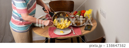 A 53-year-old woman prepares dinner at a round wooden table in her kitchen. She pours food from a colander onto a plate while arranging utensils and other items, banner 138647382