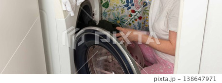 A woman takes clean clothes out of a washing machine in a laundry room. She is focused on her task, surrounded by a colorful wall. It is daytime and chores are being done, banner 138647971