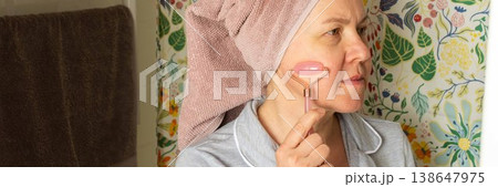 A 50 year old woman applies a facial roller to her face while standing in a bright bathroom. She has a towel wrapped around her hair and wears comfortable clothes, banner 138647975