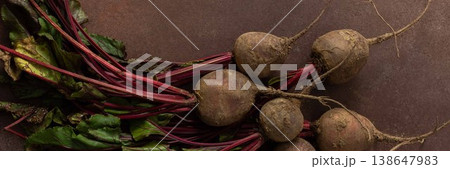 Beets with green leaves rest on a brown surface in a kitchen. The setting is warm and hints at fresh produce ready for cooking or eating, banner 138647983