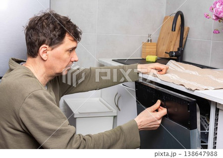 A man stands in his kitchen taking clean dishes out of the dishwasher. He focuses on the process of unloading the clean dishes and organizing them for use A man stands in his kitchen taking clean dishes out of the dishwasher. He focuses on the process of unloading the clean dishes and organizing them for use 138647988