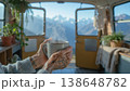 A person holds a warm cup of coffee while sitting in a plant-filled room. Large windows show a mountain vista. The sun shines through as snow tops shine in the distance. 138648782