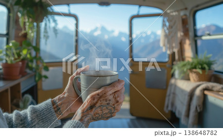 A person holds a warm cup of coffee while sitting in a plant-filled room. Large windows show a mountain vista. The sun shines through as snow tops shine in the distance. 138648782
