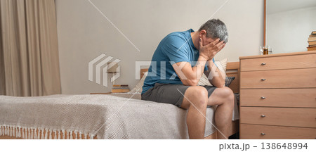 Man sitting on bed, covering face with hands showing signs of distress, loss, and mental health issues. Mental health of middle-aged people, midlife crisis 138648994