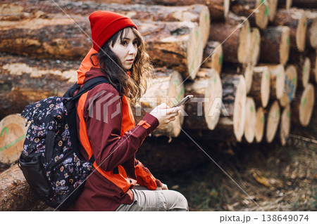 Woman with backpack sits on log while holding smartphone in front of stacked timber. hiking navigation app, mountain gps tracking, digital map outdoors, traveler using smartphone. Woman with backpack sits on log while holding smartphone in front of stacked timber. hiking navigation app, mountain gps tracking, digital map outdoors, traveler using smartphone. 138649074