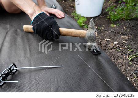 A person wearing boots and gloves is kneeling on the ground on a flower bed installing black weed fabric using galvanized steel pegs and a mallet. Concept image for DIY landscaping, weed protection. 138649135